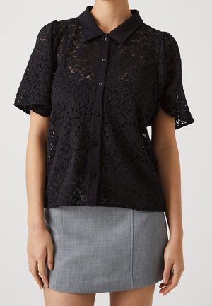 Schwarze Spitzenbluse mit kragenähnlichem Ausschnitt, kurzen Puffärmeln und Knopfverschluss. Getragen mit einem grauen, taillierten Rock. Detailliertes florales Muster.