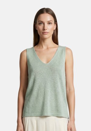 Donna con capelli castani lisci, che indossa un top a maglia verde chiaro senza maniche con scollo a V e una gonna avorio, rivolta in avanti su uno sfondo semplice.