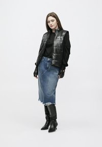 Νεαρή γυναίκα που φοράει μαύρο μπουφάν χωρίς μανίκια, μαύρη μπλούζα με μακριά μανίκια, γάντια χωρίς δάχτυλα, σκιστή τζιν φούστα και μαύρες μπότες μέχρι το γόνατο, σε ουδέτερη στάση.