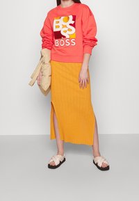 Felpa corallo con grafica "BOSS", abbinata a una gonna gialla a coste al ginocchio con spacco laterale. Sandali beige a ciabatta con cinturini.