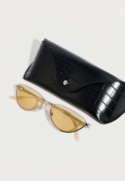 Γυαλιά ηλίου με σχήμα γάτας, με κίτρινους φακούς και λεπτά μεταλλικά πλαίσια δίπλα σε μαύρη θήκη γυαλιών με υφή και σχέδιο κροκόδειλου.