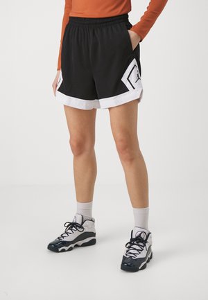 Personne portant un short de basketball noir et blanc, des chaussettes blanches et des chaussures de sport noires et blanches, debout avec une main dans la poche.