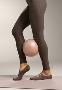 Ruskeat leggingsit, joissa sileä pinta, pitäen vaaleanpunaista jumppapalloa jalkojensa välissä. Vaaleanruskeat jalkineet, joissa ristikkäiset hihnat ja pitoiset pohjat.