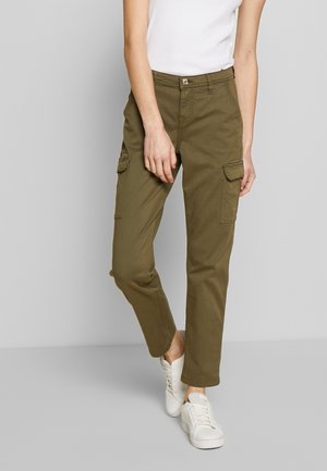 Osoba ubrana w oliwkowe spodnie typu cargo z kieszeniami z patkami oraz białe trampki, stojąca na tle jednolitego szarego tła.