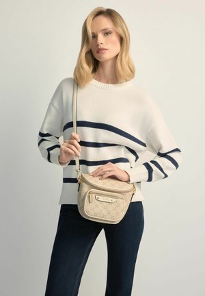 Młoda kobieta w białym swetrze w granatowe paski i ciemnych dżinsach, trzymająca beżową torebkę na ramię z wzorem i złotymi akcentami.