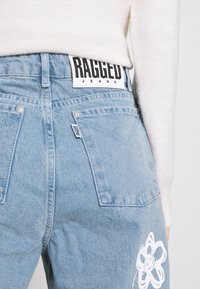 Jeans in denim blu chiaro con un motivo floreale bianco sulla gamba sinistra, dotati di un'etichetta "RAGGED JEANS" in bianco e nero sulla vita.
