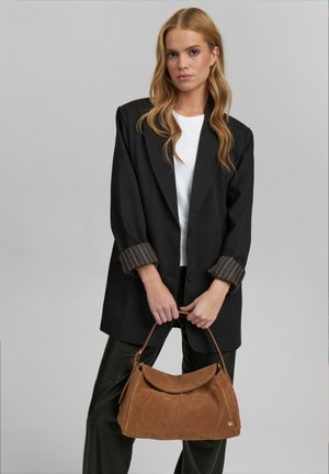 Blazer oversize nero con polsini a righe, top bianco e pantaloni scuri. Tiene in mano una borsa in suede marrone con un design curvo e morbido e manico rotondo.