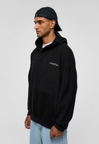 Haut à capuche noir avec une finition texturée, présentant une poche avant et une broderie blanche "Eternity Project" sur la poitrine. Associé à un jean bleu clair.