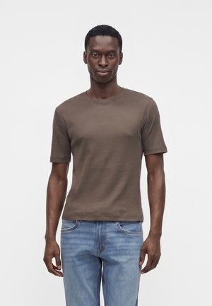 Homme debout, faisant face, portant une chemise marron à manches courtes et un jean bleu clair sur un fond blanc uni.
