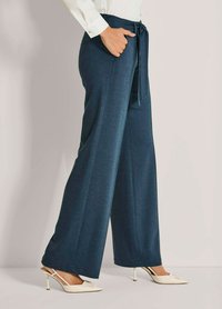 Vrouw in marineblauwe wijde broek en offwhite hoge slingback hakken, met haar hand in de broekzak.