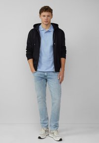 Črna zip zadrgo jutranja jakna, svetlo modra polo majica in svetlo modri kavbojke. Teniske z sivo-belim akcentom. Preprost, vsakdanji outfit.