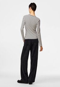 Oseba, obrnjena stran, oblečena v belo-črto dolgo majico z dolgimi rokavi, črne široke hlače in črne čevlje na enobarvnem ozadju.