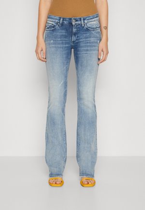 Osoba nosząca wyblakłe niebieskie jeansy bootcut, brązową bluzkę, żółte sandały z odkrytymi palcami, stojąca na jednolitym białym tle.