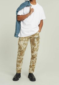 Khaki kamuflażowe spodnie cargo z wieloma kieszeniami, zestawione z gładką białą koszulką i dżinsową kurtką oraz czarnymi butami.