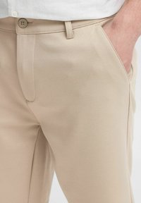 Pantalons beiges ajustés en tissu doux et texturé. Dotés d'une fermeture à bouton et d'une poche latérale, affichant des lignes épurées et un profil slim.