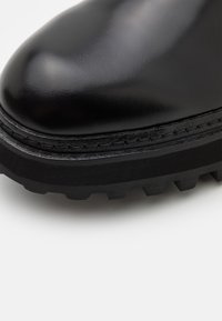 Botte en cuir noir avec une surface lisse et brillante. Elle présente une semelle noire épaisse et texturée avec une bande de roulement robuste et des détails de coutures visibles.