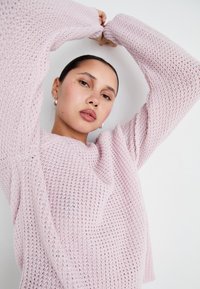 Heled roosa soonikko sweater lahtise, tekstuuriga disainiga ja laia varrukatega, mille kannab tumepealine inimene, kelle juuksed on tagasi seotud ja käed üles tõstetud.