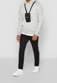 Sudadera gris con bolsillo frontal, bolsa bandolera negra, pantalones negros y zapatillas blancas. Diseño simple con acentos mínimos.