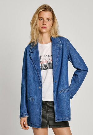 Blazer in denim blu con colletto a punta, due tasche frontali e chiusura con un solo bottone, indossato sopra una t-shirt grafica bianca.
