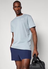 Světle modrá košile s krátkým rukávem a kulatým výstřihem, s logem Under Armour, v kombinaci s námořnickými šortkami a černou sportovní taškou.