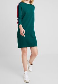 Grünes Kleid mit rundem Ausschnitt und drei Viertelärmeln, das drei horizontale Streifen in Rot, Weiß und Marineblau an den Ärmeln aufweist.