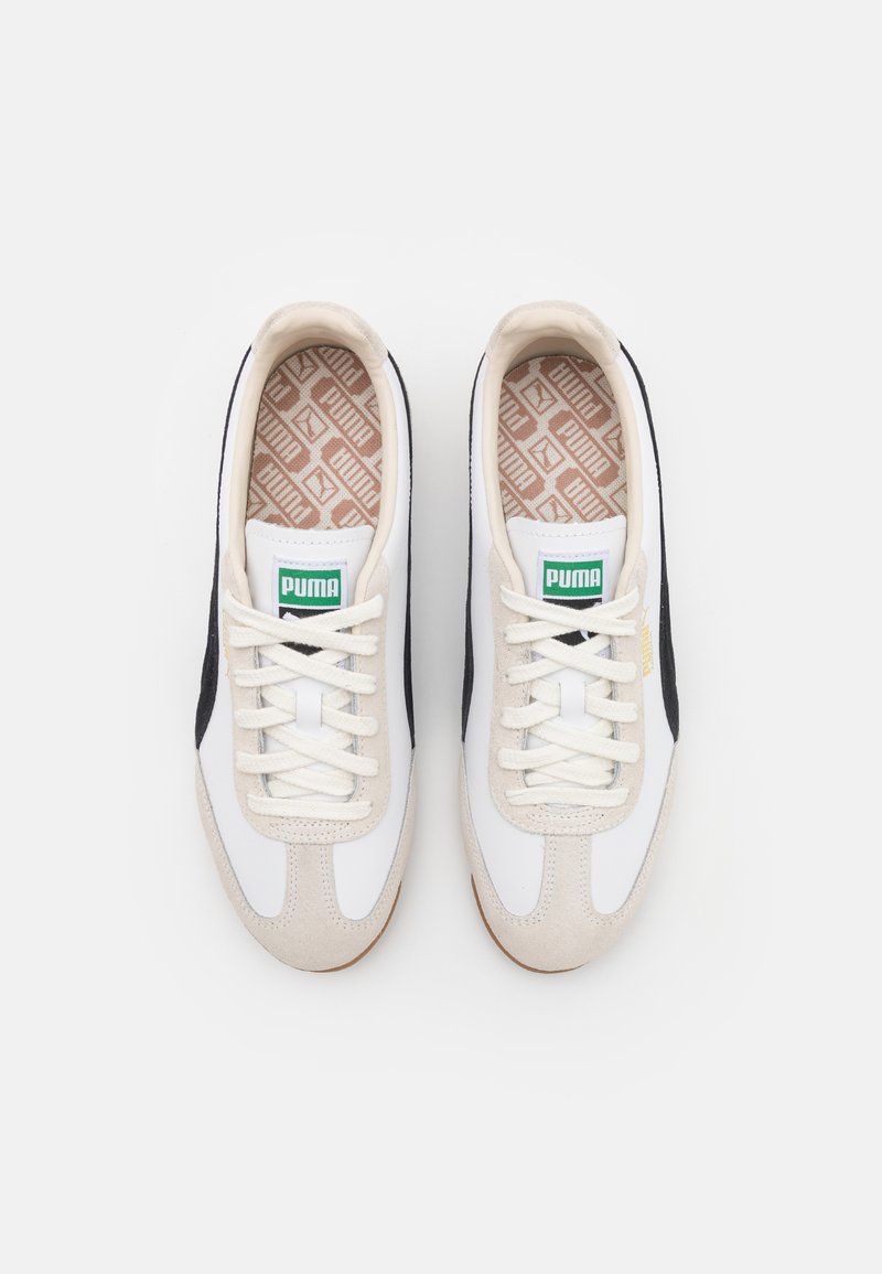 Baskets blanches et noires avec des accents en suède beige, dotées d'une semelle intérieure à motif, de lacets ronds et d'un logo Puma vert sur la languette.