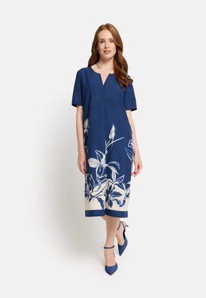 Frau trägt ein marineblaues Midikleid mit weißem Blumenmuster am Saum, kurzen Ärmeln und passenden blauen spitzen Schuhen.