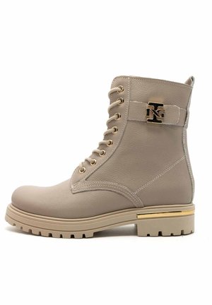 Bottine beige à lacets avec des accents en métal doré, une semelle robuste et du cuir texturé. Présente un détail de logo sur la sangle.