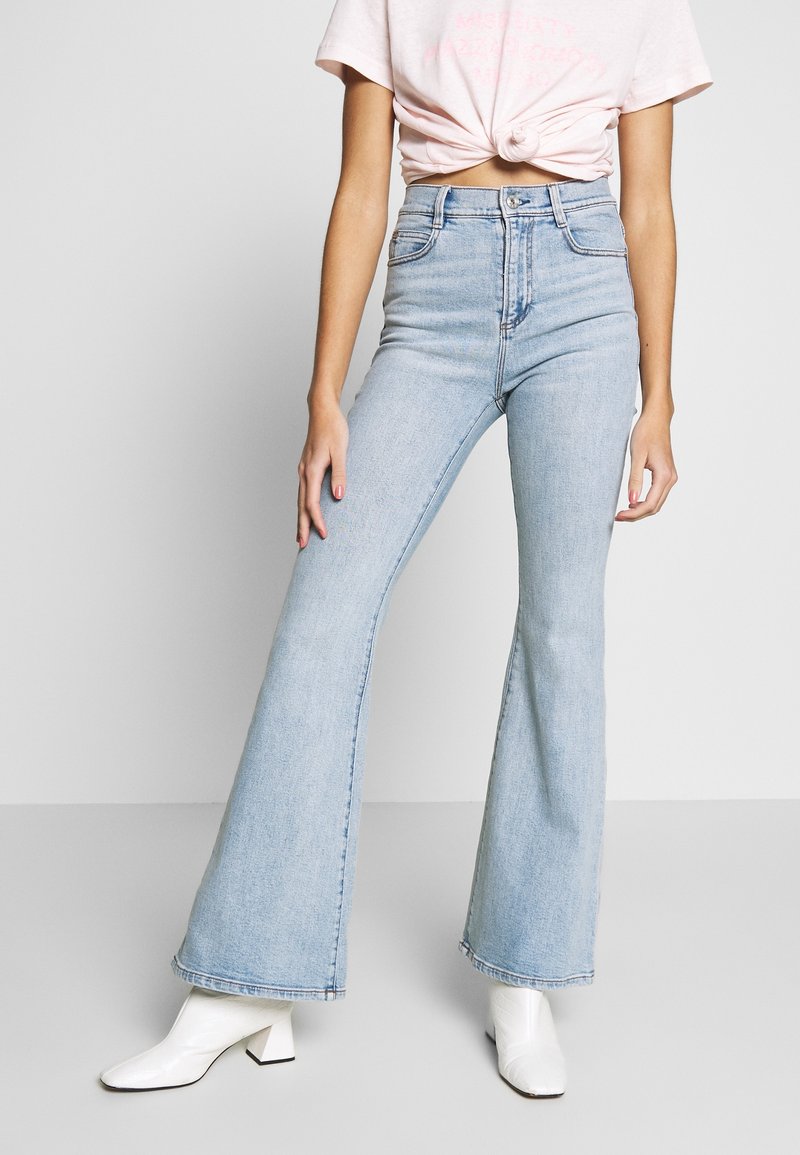 Personne portant un jean flare taille haute bleu clair, un t-shirt court noué rose pâle et des bottines blanches à talons sur un fond uni.