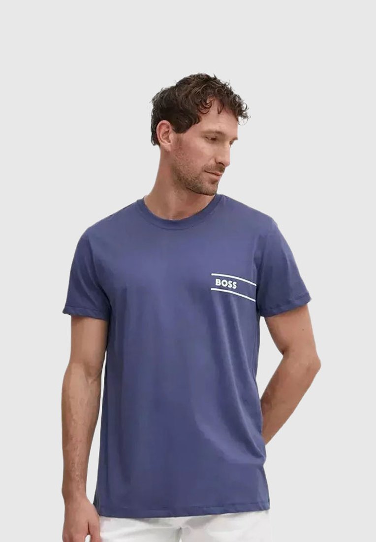 Homme portant un t-shirt bleu uni avec le texte "BOSS" sur le côté gauche de la poitrine, regardant vers le bas et sur le côté avec une main derrière le dos.