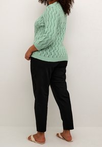 Pull tricoté vert menthe avec un motif ondulé, manches trois-quarts. Porté avec un pantalon noir resserré et des sandales de couleur claire.