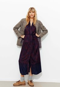 Robe longue et boutonnée en bleu marine foncé avec des motifs floraux bordeaux, associée à un blazer à carreaux. Des chaussures marron à lacets et des chaussettes grises complètent le look.