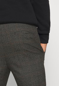 Tweed broek in donkergrijs met een ruitpatroon, voorzien van subtiele oranje accenten. Zijkant zakdetails zichtbaar, gedragen met een zwarte trui.