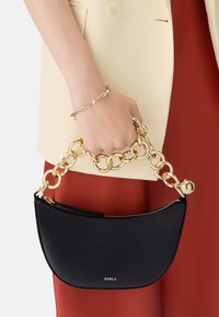 Czarna skórzana torebka na ramię z złotym łańcuchem. O zaokrąglonym kształcie, z zamkiem błyskawicznym i logo "Furla". Modelka nosi beżowy top i czerwoną spódnicę.