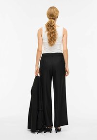 Črne široke hlače z visokim pasom in gladko teksturo, kombinirane s prilagojeno belo majico brez rokavov. Črni blazer je prevržen čez eno roko.