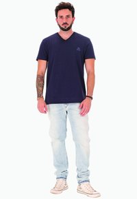 Uomo con barba e braccio sinistro tatuato, che indossa una maglietta blu marine, jeans azzurri, scarpe da ginnastica bianche e si trova contro uno sfondo bianco.