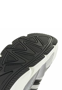Zapatilla deportiva con una suela exterior de goma negra texturizada, que presenta patrones en zigzag y detalles en blanco; parte superior en gris claro con detalles de malla.