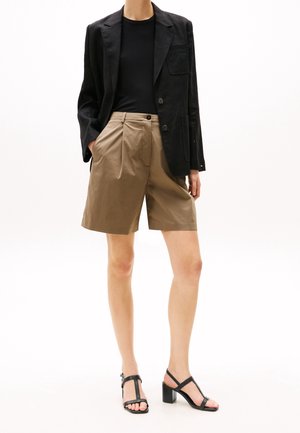 Blazer noir sur un haut noir ajusté, associé à des shorts plissés kaki et des sandales à talons noires. Textures lisses et coupe ajustée.