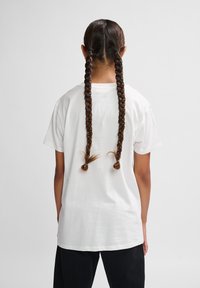 Hvid bomulds t-shirt med korte ærmer, set fra ryggen. To lange brune fletninger hænger ned med små løkker i enderne.