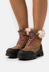 Bottes imperméables couleur fauve avec une semelle en caoutchouc noir, un manchon en fausse fourrure et des lacets multicolores. Dotées de détails de couture et d'un design à bout arrondi.