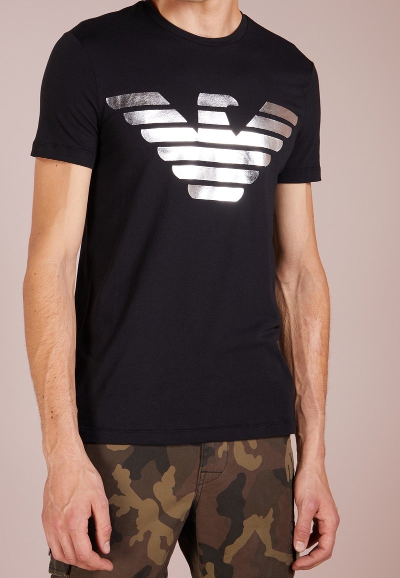Svart bomullströja med en metallisk silverörnslogotyp, korta ärmar och klassisk rund halsringning, i kombination med camouflagemönstrade byxor.