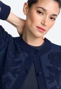 Cardigan blu navy con ricamo floreale, collo e polsini a coste e chiusura con bottoni. Texture morbida con dettagli di motivo intricati.