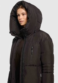 Veste matelassée noire avec une grande capuche, une fermeture éclair devant et des poches latérales. Le matériau semble lisse avec des sections matelassées et une finition brillante.