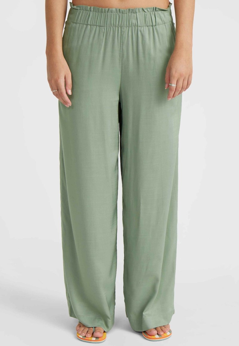 Pantalon ample vert clair avec taille élastique porté par une personne debout pieds nus sur des sandales blanches et jaunes.