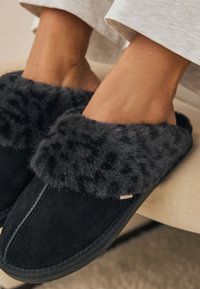 Les chaussons en daim noir sont dotés d'un manchon en fausse fourrure gris foncé et moelleux. Le modèle sans lacets présente des détails de surpiqûres et une semelle plate en caoutchouc.