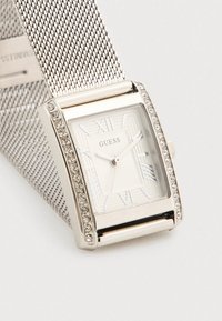 Ασημένιο ορθογώνιο ρολόι Guess με λευκό καντράν, ρωμαϊκούς αριθμούς, κρύσταλλα διακοσμητικά στη στεφάνη και μεταλλικό πλεκτό λουρί.