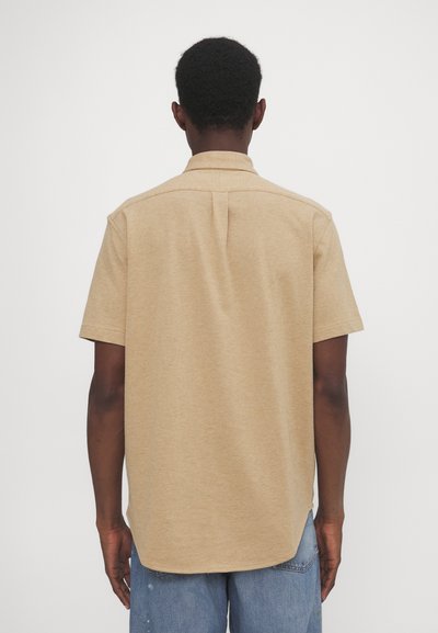 Camisa de manga curta, com gola, em tecido bege claro. Apresenta uma costura de yugo nas costas e uma bainha suavemente curva. Combinada com jeans de ganga azuis.