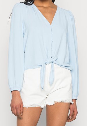 Kvinde iført en lyseblå langærmet bluse med knapper, bundet ved livet, og hvide frynsede denimshorts, vist fra skuldre til midt på låret.
