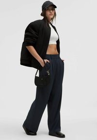 Black oversized bomber jacket, white crop top, navy wide-leg trousers, black flat shoes, small black handbag with gold hardware, and a black cap.