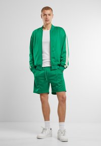 Chándal verde de tejido suave, con una chaqueta estilo bomber con rayas crema y pantalones cortos a juego. Combinado con zapatillas blancas.